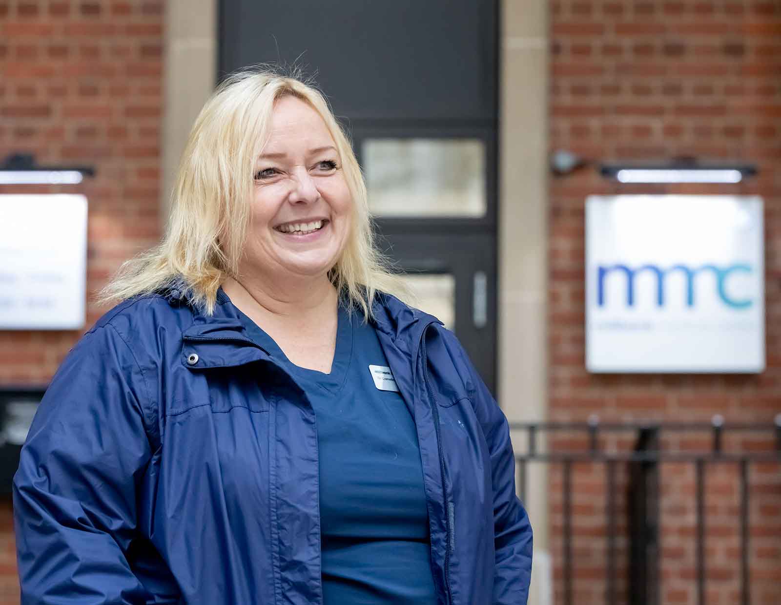 Landscape image of Dr Connie Jungen-Minton wearing a blue jacket over her scrubs, smiling outside of her practice.