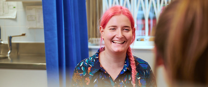Lady with pink hair smiling at another lady sitting in front of her. Both women are in a GP practice. 