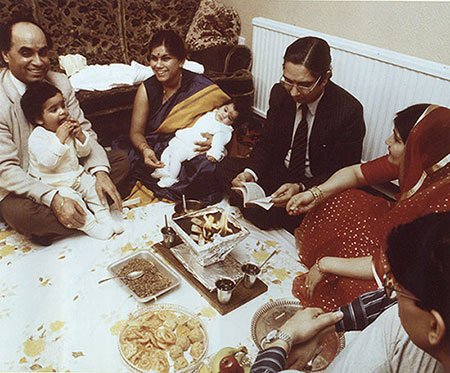 Image of Shiv Pande and his family. Shiv is holding a boy, the lady next to him a baby. There is a gentlemen wearing a suit with glasses and reading a book. Next to him is a woman wearing a red saree and next to her is a man sitting crossed legged. There are indian sweets in a tray on a white sheet covering the carpet, and there is a small holy ritual going on. 
