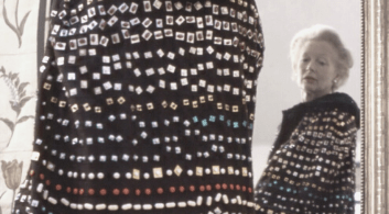 A woman with white hair wearing a bejewelled jumper looks at herself in the mirror.