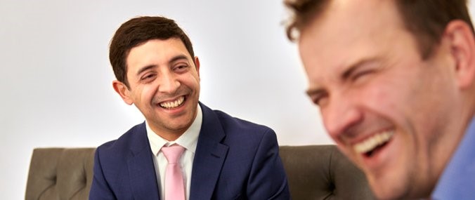 A man with dark, cropped hair looks at a laughing colleague close to the camera. 