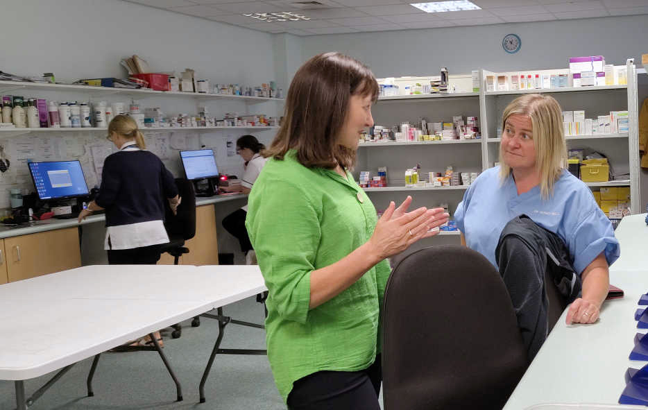 Two women having a discussion in a hospital pharmacy. One wears a green top and gestures whilst speaking, the other wears blue scrubs and listens. Medicine shelves and pharmacy staff working at computers are visible in the background.