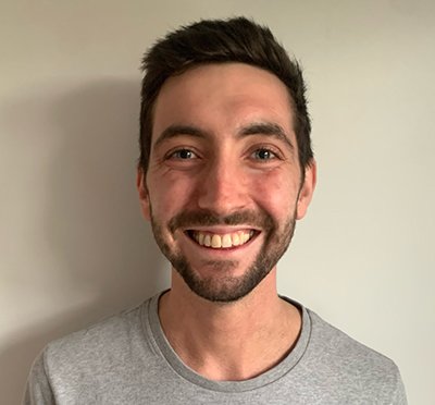 Dr Tom France smiles at the camera while standing in front of a cream wall.