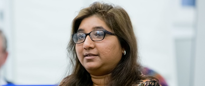 A woman in glasses and with long hair, speaking at an event while sitting in the audience