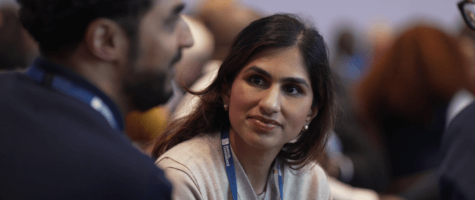 A smiling woman with dark hair looks at a talking colleague. 