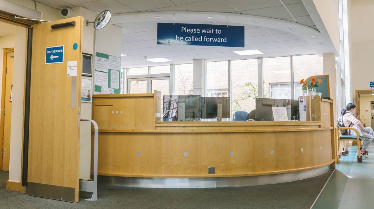 A reception desk at a doctors' clinic, with a sign saying "Please wait to be called forward" hanging overhead
