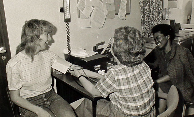 Photograph from the 1980s showing Dr. Doreen Vernon measuring a patient's blood pressure during a World Health Day event, with Dr. Pro Torkington observing.