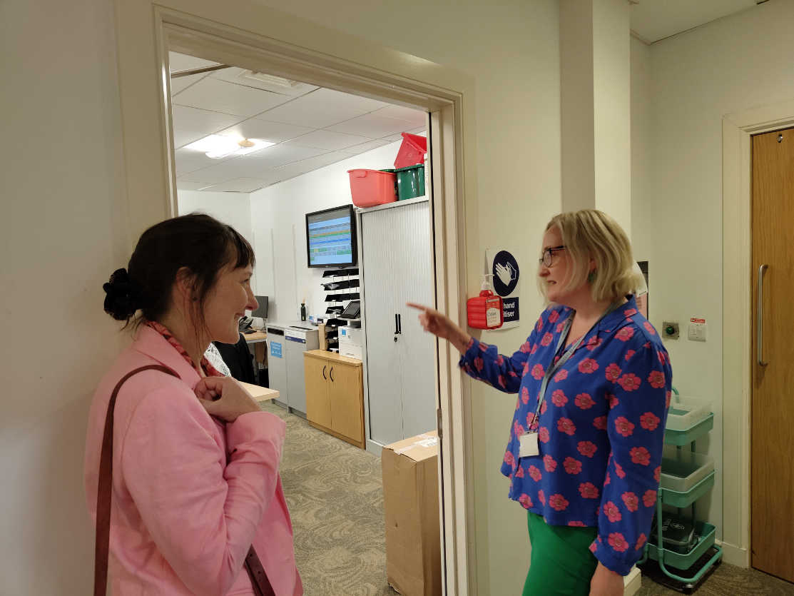 Two NHS staff members chatting in a hospital corridor. One woman wears pink scrubs, the other wears blue floral scrubs and is gesturing whilst speaking. Medical equipment and storage are visible in the background.
