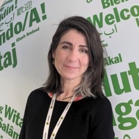 Dr Ayla Cosh stands in front of a white wall designed with large green text. 