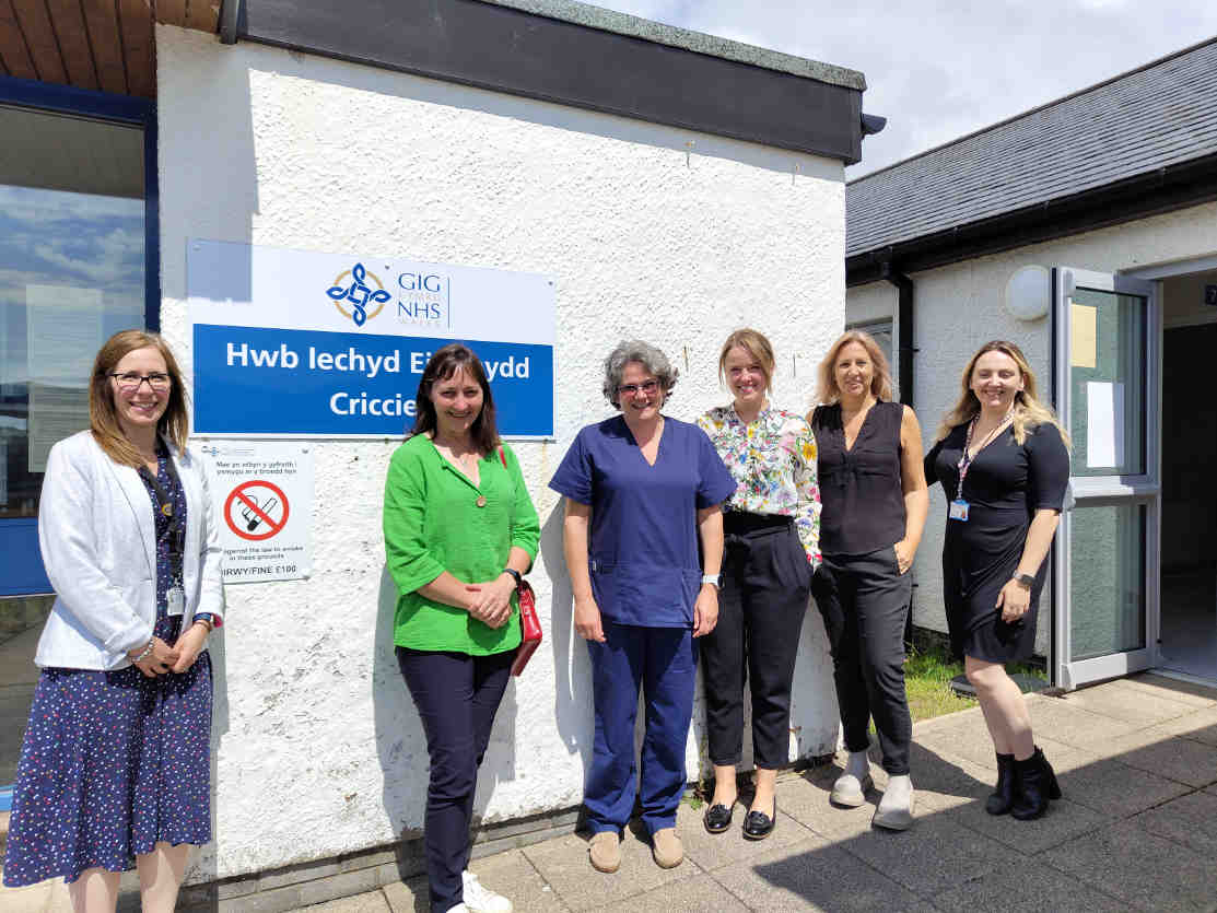 Six women posing outside a Welsh NHS health centre with bilingual signage reading 'Hwb Iechyd Brynmawr' and 'GIG NHS' logo. The group includes healthcare staff in scrubs and professional attire, standing in front of a modern medical building.