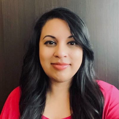 A square headshot image of Dr Mehwish Sharif wearing a bright pink jumper standing beside a dark wall. 