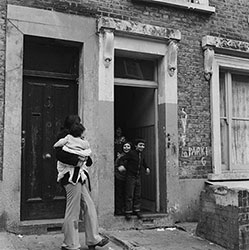 Black-and-white photo of a woman carrying a child outside a rundown building, with three smiling children at the doorway.
Family of squatters, Camden, 1973 (Henry Grant/London Museum)