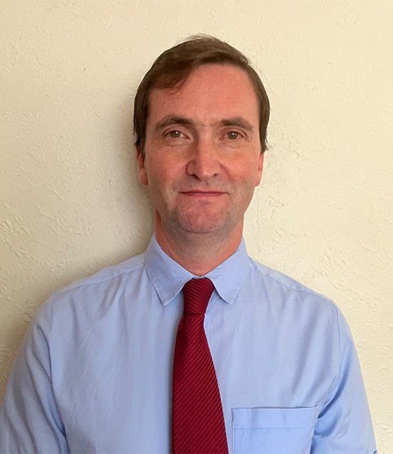 An image of Dr Chris Provan standing in front of an off-white wall wearing a light blue shirt and red tie.