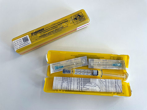 A yellow naloxone kit opened to show its contents, including a prefilled syringe, packaged needles, and instruction leaflet, with the lid and label of the case visible beside it.