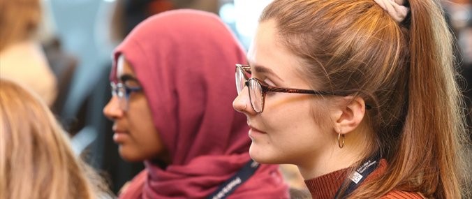 Two women in the foreground listening attentively at a professional event, with one wearing glasses and a ponytail, and another wearing a burgundy hijab.