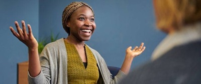 A lady wearing a dark yellow top, with a grey cardigan over it, her arms are in the air. She is smiling with a colleague sitting in front of her.