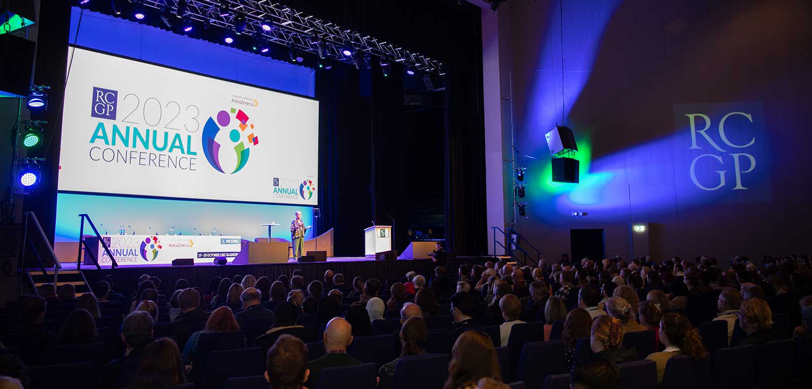 Wide shot of an audience at RCGP conference with the conference logo on a big screen on-stage