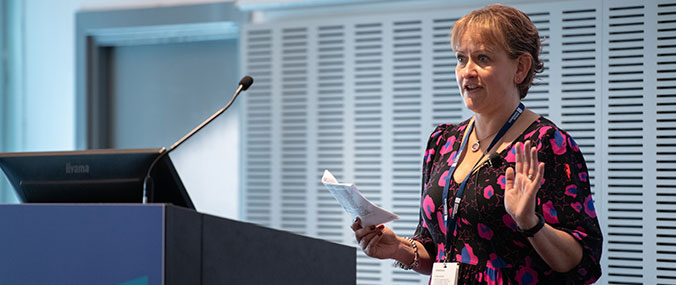 A woman with reddish-brown hair delivers a presentation at a modern lectern, wearing a vibrant floral blouse and ID lanyard. She holds papers whilst gesturing, speaking into a microphone. The contemporary conference room features light blue walls with horizontal slat panelling and large windows, creating a bright professional setting.