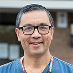 A square head and shoulders image of Dr Adrian Hayter smiling outdoors wearing blue scrubs.