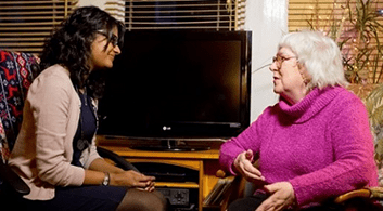 A young woman with mid-length, dark hair talks with an older woman wearing a pink jumper.