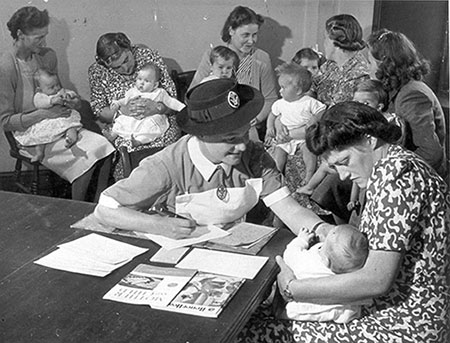 A nurse writes notes at a table while examining a baby held by a woman; other women with infants sit in the background.