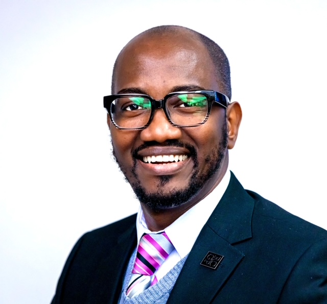 A professional headshot of Dr Uwadiae Ima Edomwonyi wearing a navy suit and pink tie.