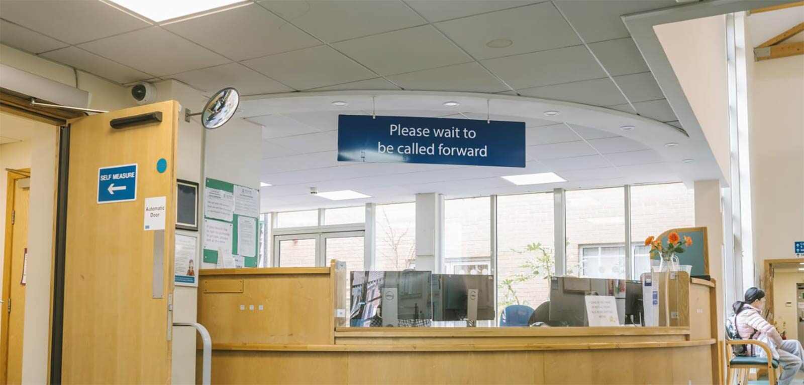 A reception desk at a doctors' clinic, with a sign saying "Please wait to be called forward" hanging overhead